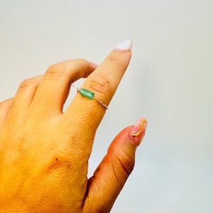 Könnte beinhalten: Ein zarter goldener Kettenring mit einem grünen Edelstein. Der Ring wird am Daumen einer Hand mit lackierten Nägeln getragen.
