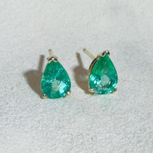 Puede incluir: Un par de pendientes de esmeralda en forma de lágrima con engastes de oro. Las piedras preciosas de color verde vibrante están facetadas, captando la luz. Los pendientes se exhiben sobre una superficie neutra, mostrando su color y diseño.