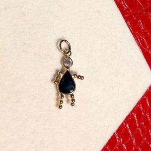 Peut inclure: Un charm doré en forme de personne. Le charm présente une pierre précieuse bleue foncée en forme de goutte d'eau pour le corps, une pierre précieuse ronde transparente pour la tête et de petites perles dorées pour les bras et les jambes. Le charm a une petite boucle en haut.