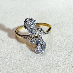 May include: A gold ring with a decorative, ornate design. The ring features two large, clear gemstones and smaller accent stones. The band is a simple gold color, and the setting is silver-toned. The ring is set against a neutral background.