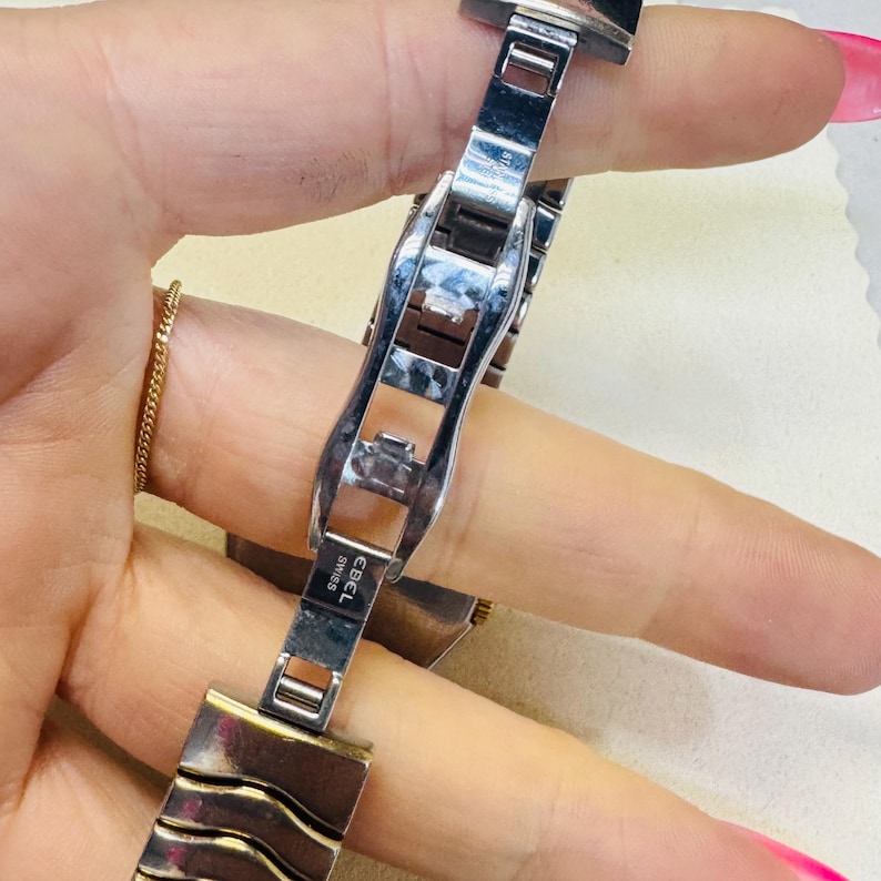 Pode incluir: Close-up de uma pulseira de rel&oacute;gio Ebel Swiss prateada e dourada. A pulseira apresenta elos retangulares com um design ondulado. O fecho est&aacute; aberto, revelando o log&oacute;tipo Ebel Swiss.