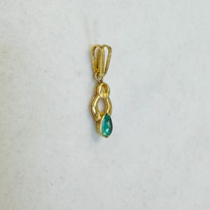 May include: A gold pendant with a teardrop-shaped, teal-colored gemstone. The pendant has a looped design at the top for a chain and a decorative, elongated shape below the loop. The gemstone is set at the bottom of the pendant.