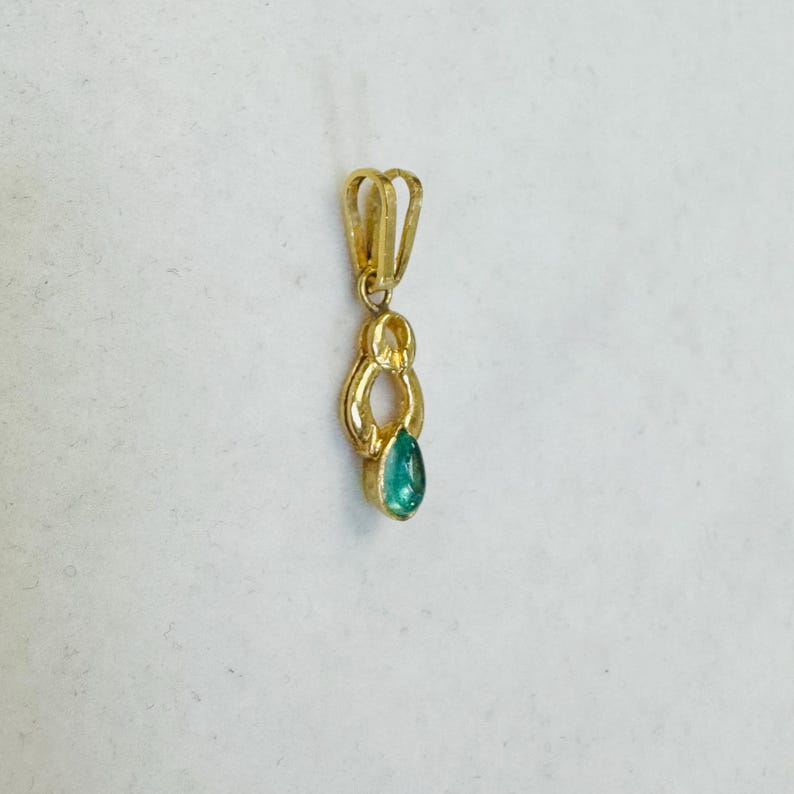May include: A gold pendant with a teardrop-shaped, teal-colored gemstone. The pendant has a looped design at the top for a chain and a decorative, elongated shape below the loop. The gemstone is set at the bottom of the pendant.