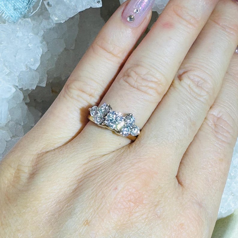 May include: A gold ring featuring a cluster of clear, sparkling gemstones. The ring's design showcases a central, larger stone surrounded by smaller stones, creating a dazzling display. The ring is set against a neutral background.