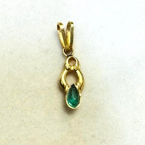 May include: A gold pendant with a teardrop-shaped emerald gemstone. The pendant has a loop at the top for a chain and a decorative design around the emerald. The emerald is a vibrant green color.