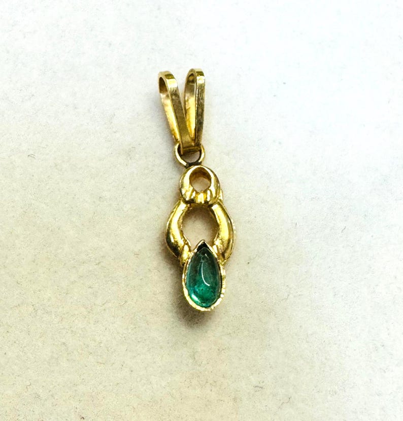 May include: A gold pendant with a teardrop-shaped emerald gemstone. The pendant has a loop at the top for a chain and a decorative design around the emerald. The emerald is a vibrant green color.