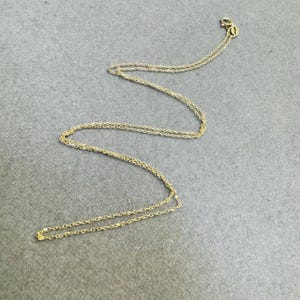 Pode incluir: Um delicado colar de corrente dourada. O colar é feito de pequenos elos de corrente. O fecho é visível no canto superior direito. O colar é exibido em uma superfície cinza.