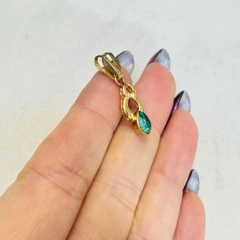 May include: A gold-colored pendant with an emerald-green gemstone. The pendant has an ornate design with a teardrop-shaped stone. The clasp is visible at the top. The jewelry is likely for a necklace or bracelet.