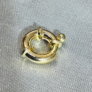 May include: A gold-toned circular spring ring clasp featuring a polished metallic finish. The jewelry component has a small lever mechanism on the side that retracts to open the ring, allowing for the attachment of chains or other jewelry items.