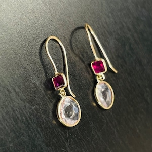 以下が含まれることがあります： ピンクと赤の宝石が付いたゴールドのドロップイヤリングのペア。イヤリングには、小さな正方形の赤い宝石と、より大きな楕円形のピンクの宝石が付いています。