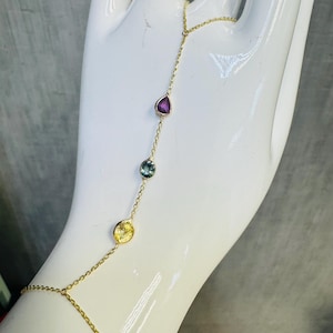May include: A delicate gold hand chain featuring a series of colorful gemstones. The chain connects a ring to a bracelet, with a teardrop-shaped purple stone, a round blue-green stone, and a round yellow stone.
