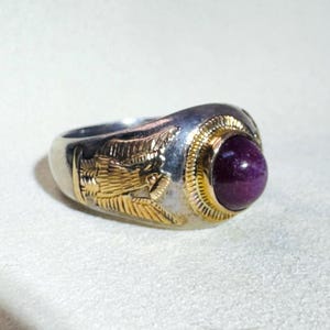 May include: A silver ring with gold accents and a large, round, deep purple gemstone. The ring features gold filigree designs on the sides and around the gemstone setting. The ring is a statement piece.