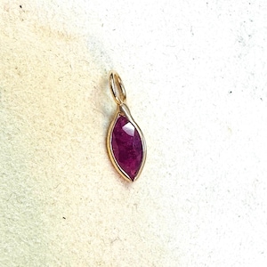 May include: A gold pendant with a marquise-shaped, faceted ruby gemstone. The ruby is a deep red color and is set in a gold bezel. The pendant has a small loop at the top for a chain.