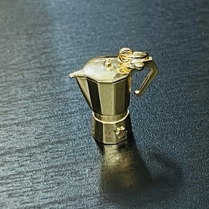 May include: A gold-coloured metal charm in the shape of a traditional Italian coffee pot, or Moka pot. The charm has a detailed design and a small loop at the top for attaching to a necklace or bracelet.