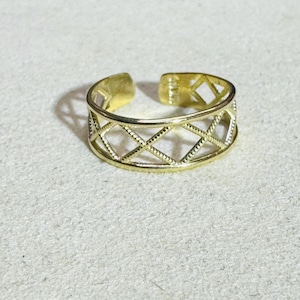 Puede incluir: Un anillo abierto de tono dorado que presenta un patrón de celosía en forma de diamante repetido. La banda tiene un acabado liso y pulido con una abertura ajustable en la parte posterior, creando un diseño geométrico continuo.