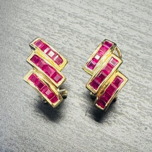 May include: A pair of gold earrings featuring a stepped, geometric design. Each earring is set with multiple square-cut, vibrant red rubies arranged in three parallel rows, creating a structured, layered appearance with a polished metal finish.