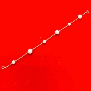 May include: A delicate gold bracelet featuring a series of white pearls of varying sizes. The pearls are strung along a fine gold chain, creating a minimalist and elegant design. The bracelet is displayed against a vibrant red background.