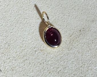 May include: A gold pendant with a deep red, oval-shaped gemstone. The gemstone is set in a gold bezel, and the pendant has a small loop at the top for a chain. The pendant is displayed on a textured, off-white surface.