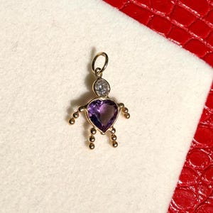 Peut inclure: Un pendentif doré en forme de personne. Le pendentif présente une pierre précieuse violette en forme de cœur pour le torse, une pierre précieuse transparente pour la tête et de petites perles dorées pour les bras et les jambes. Le pendentif est sur une surface blanche.