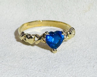 May include: A gold-colored ring with a heart-shaped, bright blue gemstone. The ring's band features a decorative design with small heart shapes. The gemstone is the focal point, set in a prong setting.