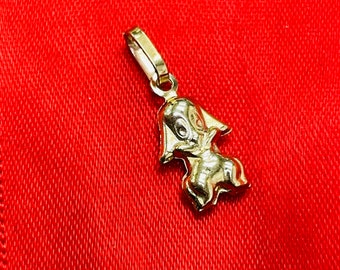 May include: A gold-colored pendant in the shape of a cartoon dog, with a loop for a chain. The pendant is set against a red fabric background. The dog has large ears and a smiling face.