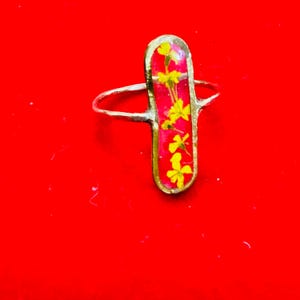 Może przedstawiać: Srebrny pierścionek z prostokątnym czerwonym i żółtym kamieniem z wciśniętymi kwiatami.