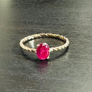 May include: A silver ring with a twisted band design. The ring features a vibrant, oval-shaped, red gemstone at the center. The ring is set against a dark background, highlighting the gemstone's color and the ring's intricate details.
