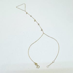 Könnte beinhalten: Zarte goldene Handkette mit kleinen, funkelnden Edelsteinen. Die Kette hat einen Ring, der um den Mittelfinger geht, verbunden mit einer Armband-Kette mit einem Verschluss. Die Edelsteine sind gleichmäßig entlang der Kette angeordnet.
