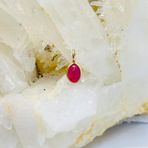 May include: A small, oval-shaped pendant with a bright pink gemstone set in a gold frame. The pendant is resting on a white, crystalline surface.