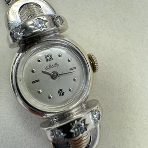 K&ouml;nnte beinhalten: Eine silberne Armbanduhr mit wei&szlig;em Zifferblatt und Diamantbesatz am Armband. Das Zifferblatt hat schwarze Zeiger und schwarze r&ouml;mische Ziffern f&uuml;r die Stunden. Auf dem Zifferblatt steht auch der Text "LE CUIlTRE".