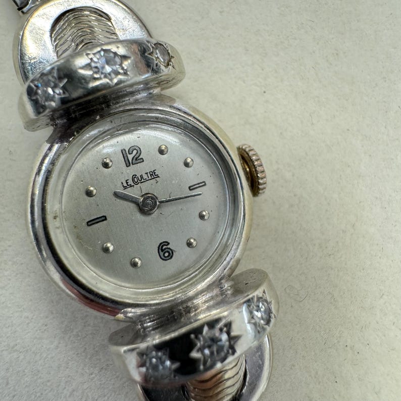 K&ouml;nnte beinhalten: Eine silberne Armbanduhr mit wei&szlig;em Zifferblatt und Diamantbesatz am Armband. Das Zifferblatt hat schwarze Zeiger und schwarze r&ouml;mische Ziffern f&uuml;r die Stunden. Auf dem Zifferblatt steht auch der Text "LE CUIlTRE".