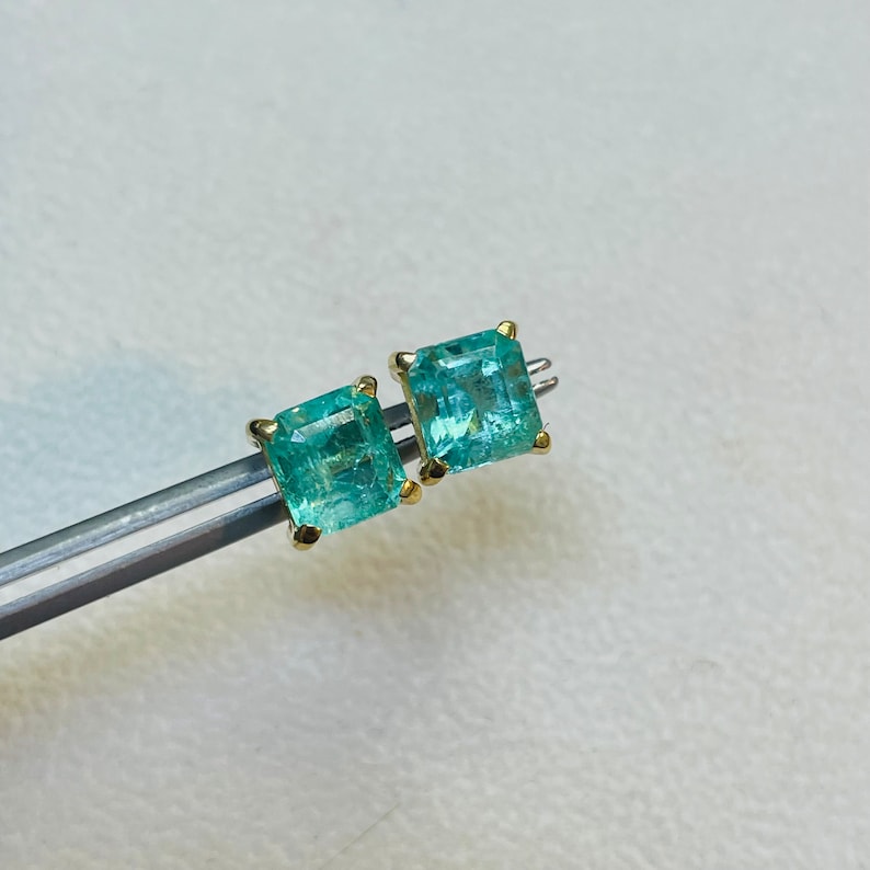 May include: A pair of emerald green gemstone earrings with gold settings. The earrings are square shaped and have a faceted cut.