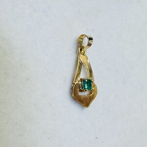 May include: A gold pendant with a rectangular emerald gemstone set in the center. The pendant has a teardrop shape with a loop at the top for a chain. The gold has a polished finish.