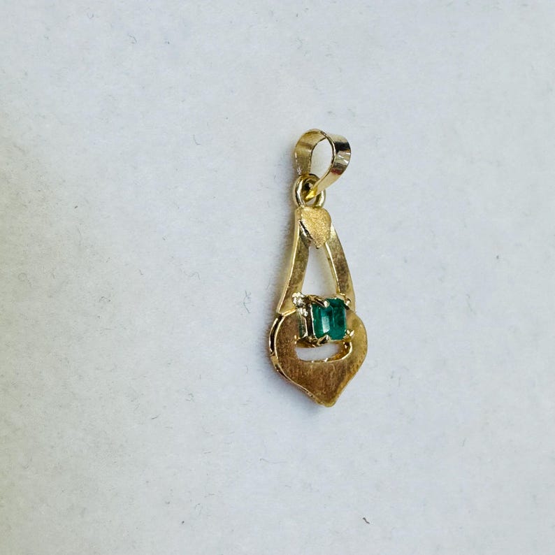 May include: A gold pendant with a rectangular emerald gemstone set in the center. The pendant has a teardrop shape with a loop at the top for a chain. The gold has a polished finish.