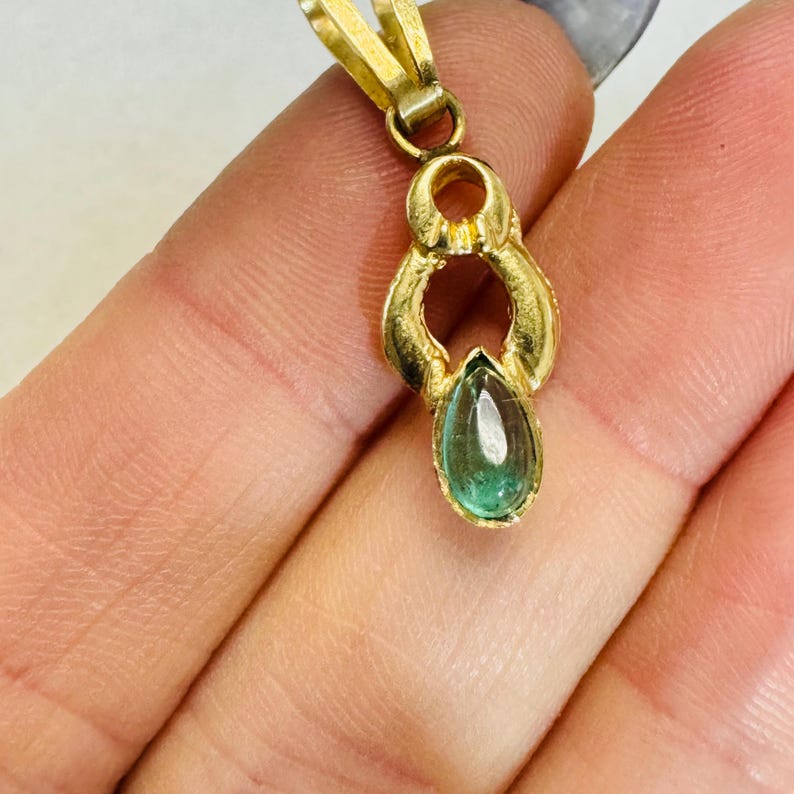 May include: A gold pendant with a teardrop-shaped, light green gemstone. The pendant has a decorative, openwork design and a small loop for a chain. The gemstone is set in a gold bezel.