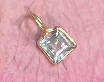 May include: A gold pendant featuring a square-cut clear gemstone set in a simple metal frame with a small circular bail at the top. The pendant is positioned against a textured pink background with visible organic veins and subtle light reflections.