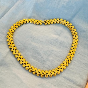 Puede incluir: Un collar dorado con un diseño de chevrón repetido. El collar está hecho de segmentos unidos, creando una joya flexible y elegante. El collar se muestra sobre una superficie azul.