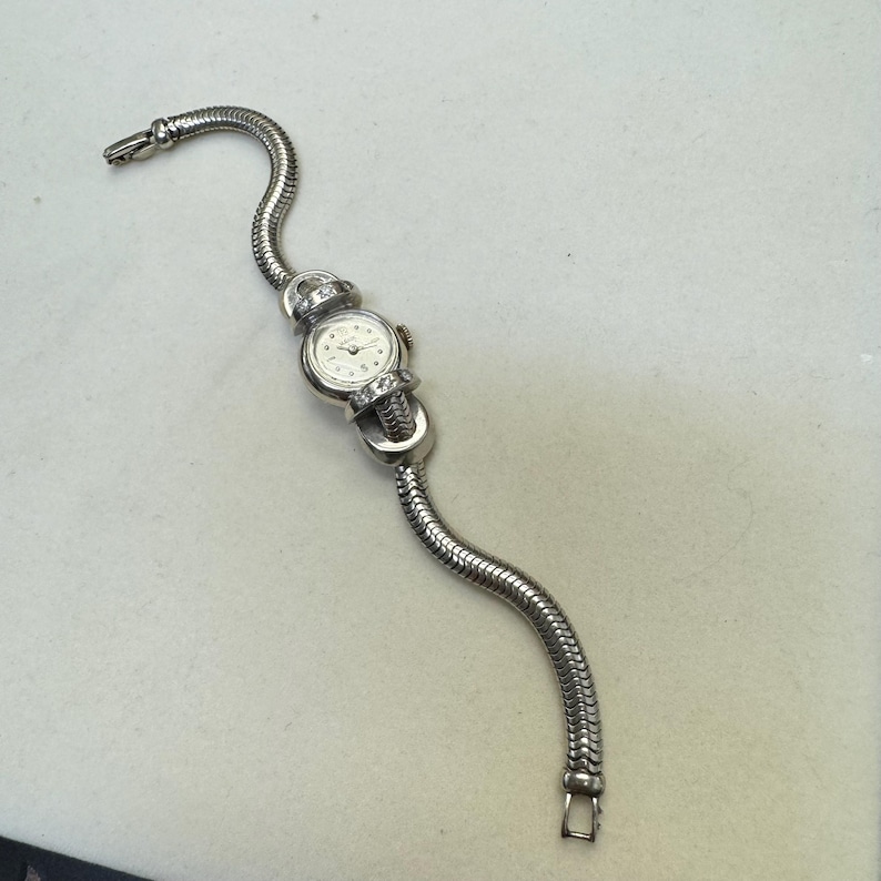 K&ouml;nnte beinhalten: Eine silberne Armbanduhr mit wei&szlig;em Zifferblatt und Diamantbesatz am Armband. Die Uhr hat ein Armband im Schlangenketten-Stil.