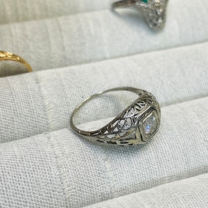 May include: A delicate filigree ring with a single, clear, square-cut gemstone set in a bezel setting.