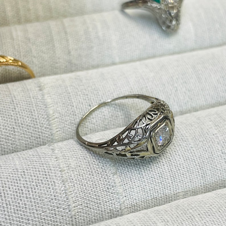 May include: A delicate filigree ring with a single, clear, square-cut gemstone set in a bezel setting.