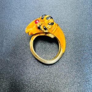 Könnte beinhalten: Ein goldfarbener Ring mit einem Widderkopf-Design. Der Widderkopf ist in Gelb, Schwarz und Rot bemalt. Der Ring hat ein silberfarbenes Band. Der Ring ist vor einem dunkelgrauen Hintergrund platziert.