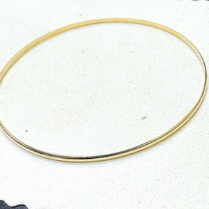 Könnte beinhalten: Ein dünnes, goldfarbenes Armreif-Armband. Das kreisförmige Armband ist schlicht und elegant, mit einer glatten, polierten Oberfläche. Der Schmuck ist vor einem weißen Hintergrund platziert, was sein minimalistisches Design hervorhebt.