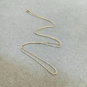 Pode incluir: Um delicado colar dourado com uma corrente fina. O colar está colocado sobre uma superfície cinza. A corrente tem um pequeno fecho na parte superior. O colar é uma joia simples e elegante.