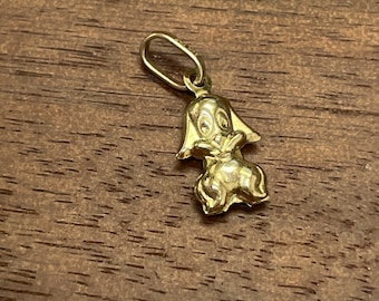 May include: A gold-colored charm in the shape of a cartoon dog. The dog is standing on its hind legs with its front paws raised. The charm has a loop at the top for attaching to a necklace or bracelet.