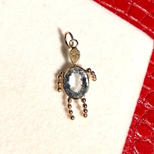 Peut inclure: Pendentif doré en forme de figurine stylisée. Le corps est une pierre précieuse ovale bleu clair, avec de petites perles dorées formant les bras et les jambes. Le pendentif est posé sur une surface blanche, avec un fond texturé rouge.