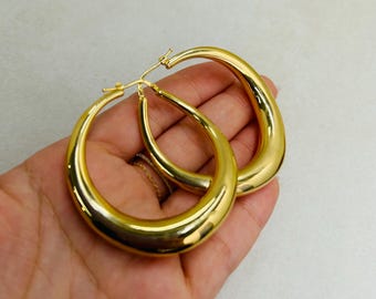 May include: A pair of gold-colored hoop earrings. The earrings have a smooth, polished surface and a teardrop shape. The earrings are held in a hand against a neutral background.