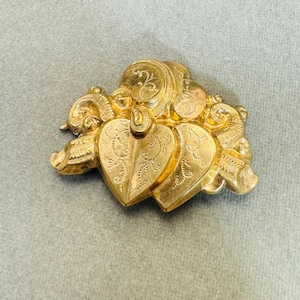 May include: A gold-toned brooch with two interlocking heart shapes, each with an intricate engraved design. The hearts are surrounded by ornate scrollwork.