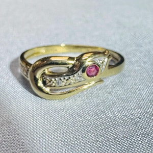 May include: A gold ring featuring a bypass-style design with a central round pink gemstone. The band includes a section accented with small, clear, sparkling stones set in a row, creating a contrast against the polished gold metal finish.