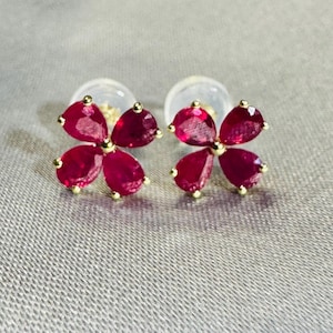 May include: A pair of stud earrings featuring four pear-shaped, deep red rubies arranged in a flower-like cluster. Each ruby is set in a gold-toned metal prong setting, with a small gold bead at the center connecting the four gemstone petals.