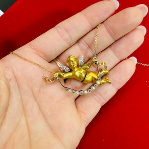 May include: A gold necklace with a cherub pendant. The cherub is holding a ribbon and has diamond accents. The necklace is adorned with pearls and diamonds.
